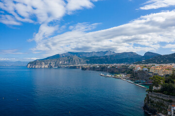 Obraz premium The dramatic cliffs and colorful buildings of Sorrento overlook deep blue Mediterranean waters, with sweeping cloud formations stretching across the sky. Yachts and boats are moored along the marina