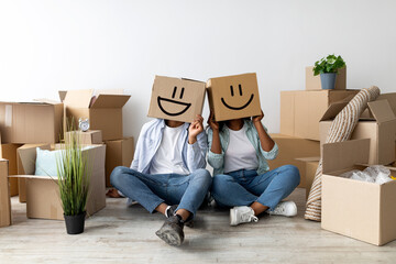 Moving and real estate concept. Black couple with cardboard boxes with smiley faces on head, sitting on floor and having fun, celebrating relocation to their own apartment