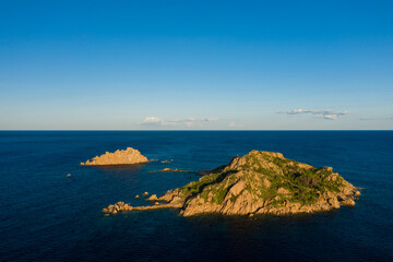 Obraz premium Two rugged islets with green vegetation rise from deep blue Mediterranean waters near Lotzorai, Sardinia. Warm golden sunlight enhances the rocky textures under a vast, clear sky.