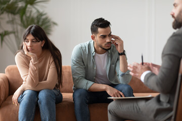 Fototapeta premium Marital Therapy Concept. Depressed stressed Middle Eastern husband and wife ignoring each other, not talking, upset millennial couple sitting apart on couch at therapist office not ready to compromise