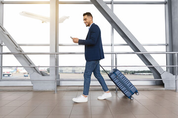Handsome arab guy using smartphone while walking with suitcase at airport terminal, young middle...