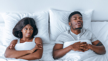 A man sleeps soundly in bed while a woman looks frustrated and annoyed. They are in a bedroom with white bedding, showcasing contrasting feelings in a shared space.
