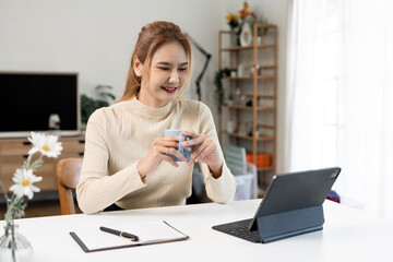 Happy Asian woman using tablet and taking notes in cozy home office. Concept of remote working,...