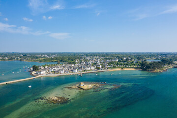 Drone view of a picturesque coastal village with white stone houses, sandy shoreline, and a small tidal islet surrounded by clear blue-green water under a bright sky in Brittany.