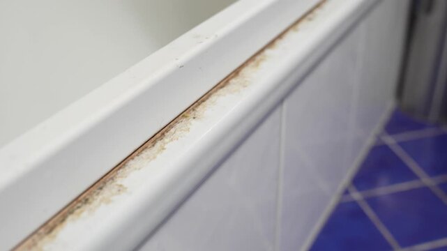 Close-up along a shower tray rim showing dirty caulk, brown staining, and peeling sealant near a corner, highlighting moisture damage and bathroom repair needs.
