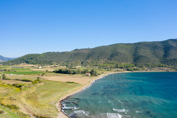 Aerial view of open farmland meeting a curving shoreline with clear blue waters, set against a backdrop of lush green hills under a bright, cloudless sky. The tranquil rural landscape features gentle