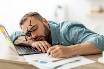 Exhausted millennial man sleeping on his office desk, next to laptop and documents, tired of...