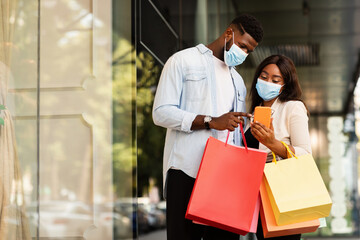 Loyalty Program, Cashback Concept. Portrait Of Happy Black Couple In Surgical Face Masks Using Smartphone Checking Bank Account After Successful Shopping In Mall During Covid Pandemic, Free Copy Space