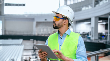 A worker in a hard hat and safety vest stands in a factory, using a tablet to monitor tasks while observing equipment. Sunlight enters through large windows.