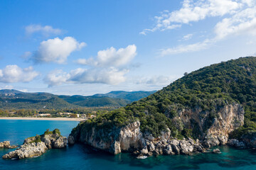 Lush green hillside rises above rugged rocky cliffs along the turquoise shoreline at Karavostasi in Epirus, Greece. Bright daylight and scattered clouds highlight the Mediterranean landscape and