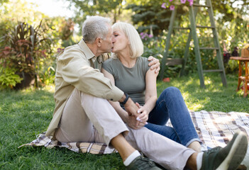 Married senior couple resting outdoors and kissing, enjoying holiday while sitting on blanket on grass in garden, free space. Happy retirement and family concept