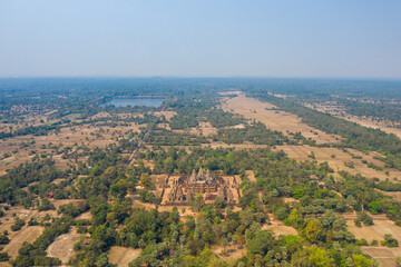 Obraz premium Expansive aerial perspective of the ancient Pre Rup temple complex surrounded by dry fields, scattered trees, and distant water reservoirs in the Angkor region of Cambodia. The scene captures the