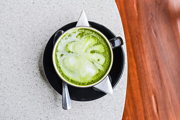 Top view of hot matcha latte in the black mug or cup. flat lay. high angle, above. spoon. stainless steel. tissue. table. grey or gray. white. green. wooden chair. copy space, empty, free, negative.