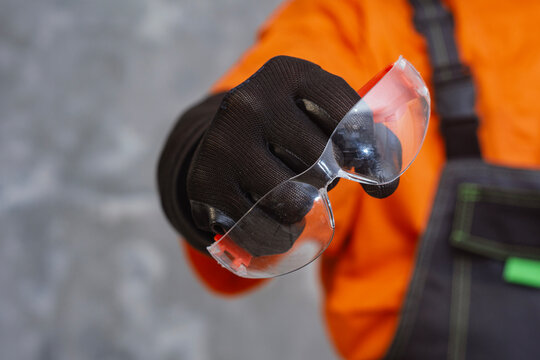 Safety glasses held in gloved hand against concrete wall. Personal protective equipment concept for construction, industrial work, repair, and workplace safety.