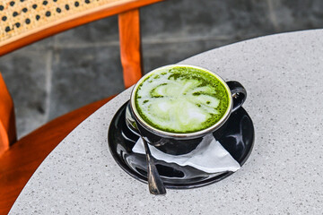 Close up view of hot matcha latte in the black mug or cup. spoon. stainless steel. tissue. table. grey or gray. white. green. wooden chair. rattan. granite table.