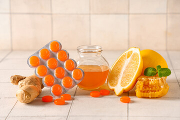 Cough drops in blister with lemon, mint leaves, ginger root and jar of honey on white tile...