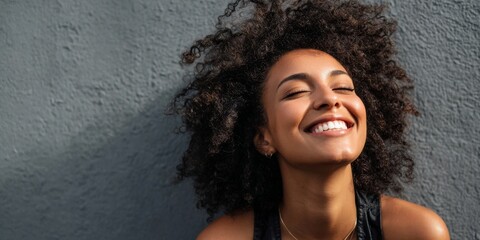 Happy woman with curly hair smiling joyfully against a textured gray wall, showcasing confidence and positivity in a vibrant portrait.