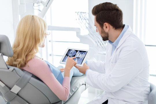 Female patient asking male dentist something, poiting at pad showing her jaws x-ray, modern dental clinic interior. Dentist and woman in dental chair discussing treatment, using digital tablet - Powered by Adobe