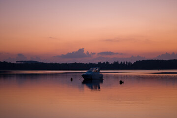 A single motorboat floats on the glassy surface of Lac des Settons at sunrise, surrounded by gentle pink and orange reflections and silhouetted forest on the distant shore.