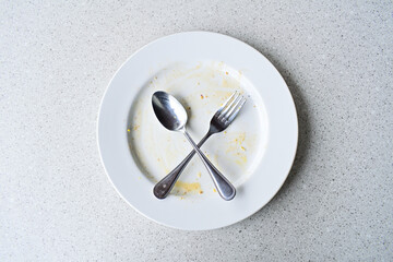Empty white plate with leftover food and utensils after a meal. Top view, high angle, above, flat lay. close up. isolated. granite table on the background. fork and spoon. stainless steel. crossed.