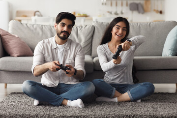Obraz premium Online video game, have fun together at home, domestic entertainment at spare time. Happy millennial arab female and guy with joysticks play on computer, on floor in living room interior, copy space