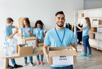 Young arab male volunteer in blue uniform joyfully holds a cardboard box labeled food donations. He is working at a charity center with friends who are preparing more donations.