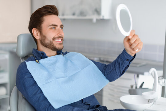Male patient looking at his beautiful healthy white smile, holding dental hand mirror, happy young man checking results of treatment at modern dentistry, using hand mirror, side view