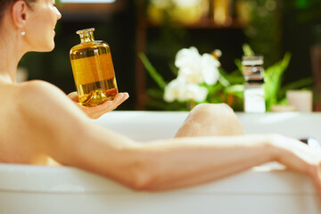 A mature woman, from a high angle, relaxes serenely in a white bathtub, holding a translucent bottle of golden bath oil. Soft light & blurred green plants create a luxurious, self-care oasis.