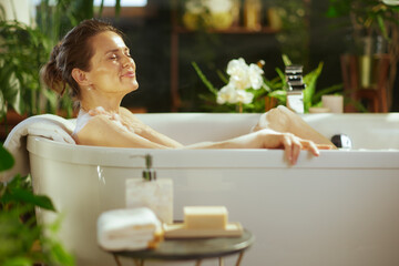 Adult woman, eyes closed, content, relaxes in white bubble bath, gently rubbing sudsy shoulder. Tranquil scene, enhanced by lush green plants & soft lighting, embodies peaceful self-care ritual.