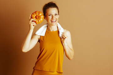 A smiling woman in orange athletic wear, with a towel, holds a small pumpkin aloft, embodying a cheerful blend of fitness, nutrition, and healthy living within the vibrant spirit of the autumn harvest