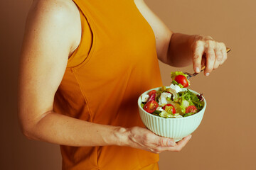 Person in yellow top holds vibrant green salad, using fork to select lettuce and cherry tomato. The image highlights conscious, healthy eating and nutritious lifestyle against simple brown backdrop