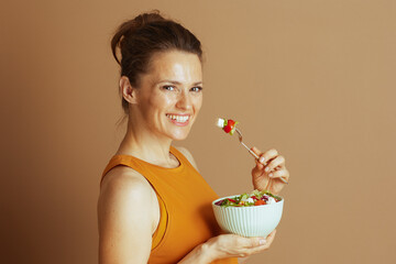 A smiling woman with a bun, wearing a mustard yellow top, looks at the camera while holding a bowl of fresh salad. She embodies a positive and healthy lifestyle against a warm brown background.