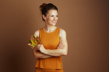A smiling middle-aged woman in a mustard yellow tank top stands with crossed arms, holding autumn leaves, looking right with a content expression, conveying autumnal serenity.