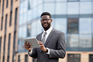 Fototapeta premium Positive african american entrepreneur using digital tablet during lunch break, walking near office center outdoors and smiling at camera, copy space