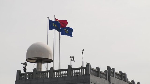 Chinese national flag and two blue maritime flags fluttering in the wind atop a stone rooftop equipped with a large white radar dome and weather monitoring sensors in Shanghai.