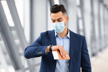Arab businessman in medical mask walking at airport terminal and checking time at wristwatch, middle eastern man holding passport with tickets and looking at watch, getting late for flight, closeup