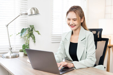 Happy Asian businesswoman in a green blazer smiling while working on a laptop at her bright home...