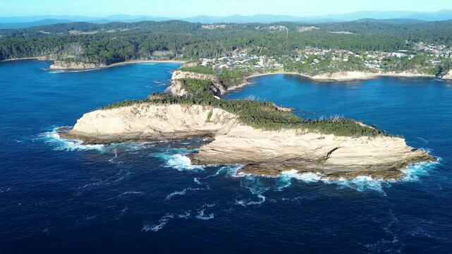 Drone aerial landscape of waves breaking on rocky ocean headland with cliffs covered in bushland trees and suburban rural town in background Malua Bay coastline South Coast Australia travel tourism
