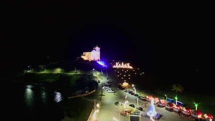 Drone Flying forward over residential houses towards the brightly lit Church of Nossa Senhora de Nazareth on the hill. Saquarema, Rio de Janeiro, Brazil. Christmas lights. 4K.