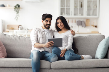 Covid-19 quarantine, online call, conference, communication in internet, watch video and new app. Cheerful young arab man and woman looking at tablet, talk with friends in living room interior