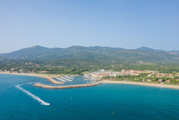 Fototapeta premium Aerial view of a marina with boats docked along a curved breakwater, bordered by turquoise water and a sandy beach, with lush green hills and mountains rising in the background under a clear blue sky.