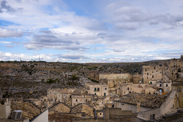 Obraz premium A panoramic view of Matera, Italy, reveals weathered stone rooftops and clustered historic buildings set against a dramatic landscape of rocky hills under a partly cloudy sky. Subtle light and natural