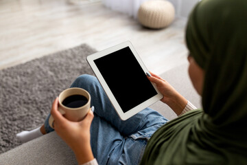 A millennial Arab woman is seated at home, sipping coffee from a cup while using a digital tablet with an empty screen.