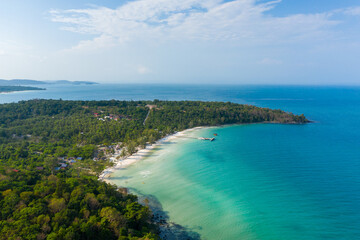 Fototapeta premium Turquoise waters meet a white sandy shoreline along a lush, forested peninsula at Coconut Beach. The scene features a wooden pier, scattered small buildings, and vibrant tropical vegetation under a