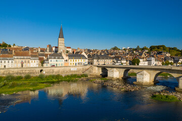 Fototapeta premium Historic stone bridge arches across the Loire river toward pastel houses and the tall abbey tower of La Charite sur Loire. Bright sunlight and a vivid blue sky highlight the town's medieval