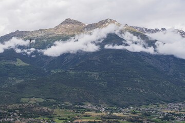 Scenic alpine landscape in Aosta Valley, Italy. Mountain village in a green valley surrounded by high peaks with snow and clouds, showing natural beauty and rural life.