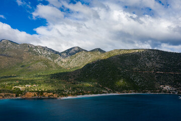 Rolling green mountains rise above a vibrant blue bay, with patches of sunlight breaking through dramatic clouds and illuminating the rugged landscape. The coastline features scattered houses and a