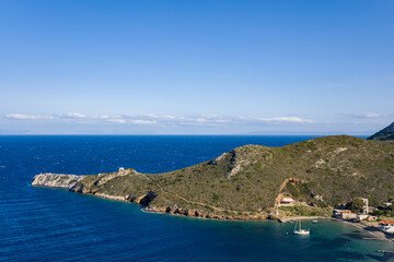 A rugged green headland extends into deep blue sea at Porto Kagio, Greece, with a sailboat anchored in a sheltered bay. The scene is bright and clear, featuring textured hills, sparkling water, and a