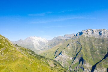 Expansive green valley flanked by rugged mountain slopes and distant sunlit summits under a clear blue sky near Gavarnie Gedre in the Pyrenees. Bright daylight reveals crisp textures and dramatic
