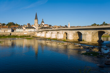 Golden sunlight highlights the stone arches of the historic bridge and the prominent abbey spire in La Charite sur Loire, with traditional houses lining the riverbank. Calm water and clear blue sky